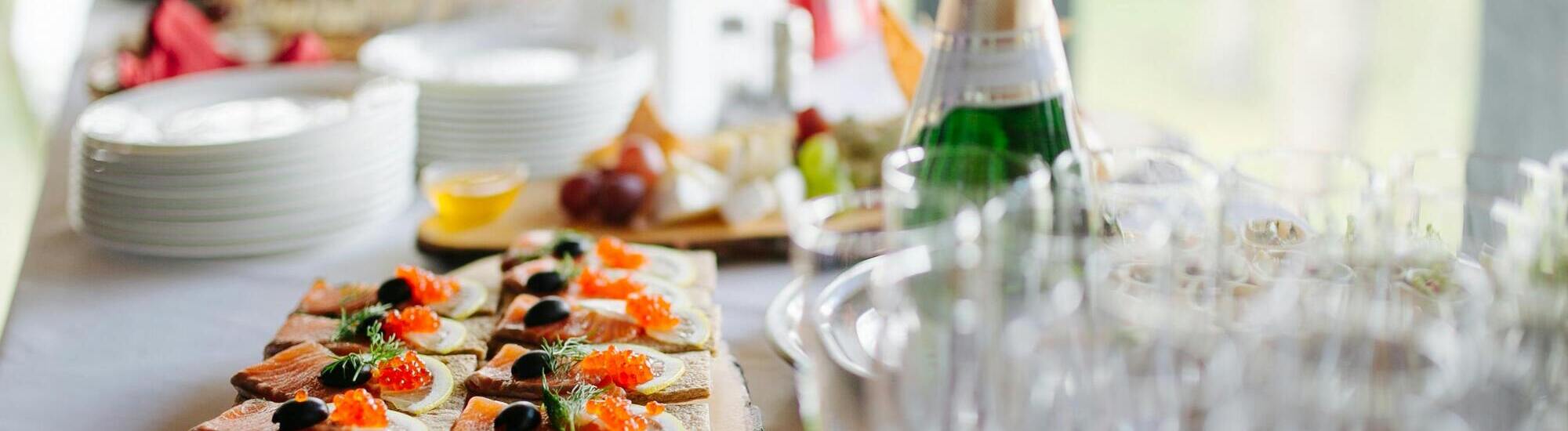 A table set with plates of appetizers topped with red caviar and herbs, a bottle of sparkling wine, empty glasses, and a cheese and fruit platter in the background.