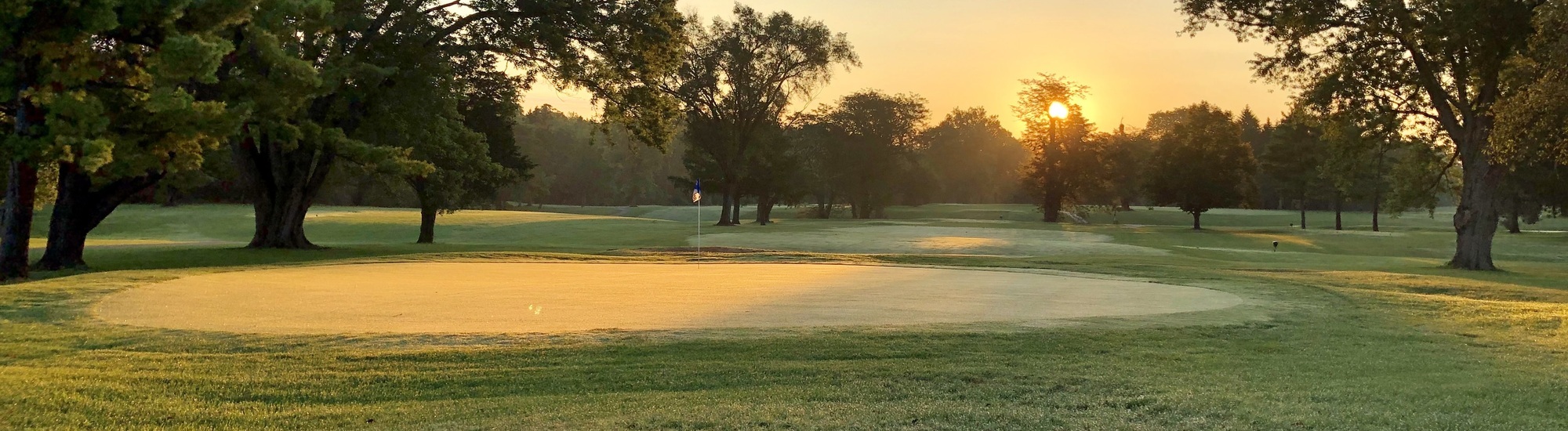 A golf course at sunrise with sunlight streaming through trees, casting long shadows and a warm glow over the green and dew-covered grass.