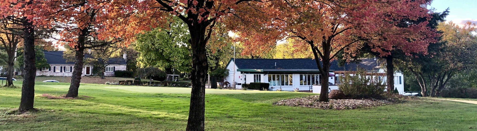 A white house with large windows sits on a green lawn, framed by trees with vibrant red and orange autumn leaves. The scene is peaceful and well-maintained, capturing the beauty of fall.