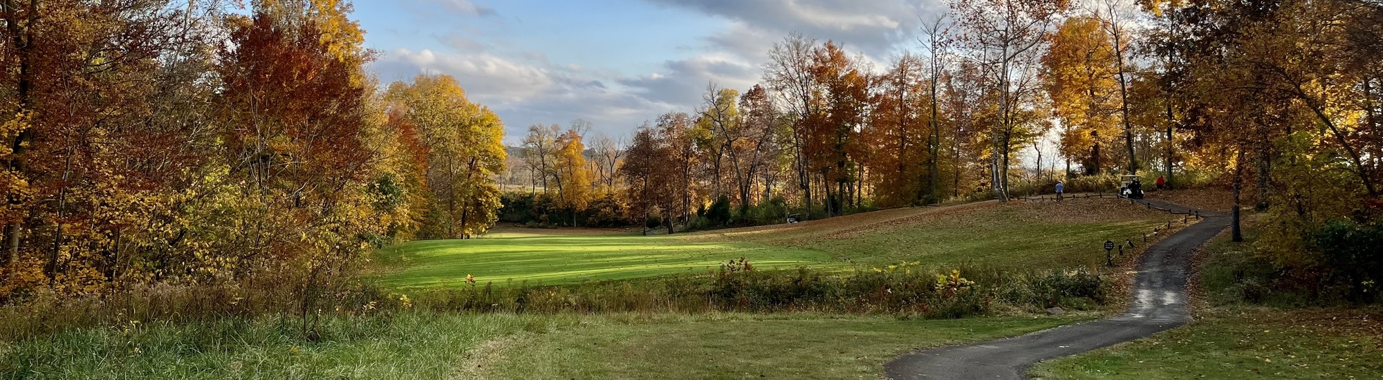A paved path curves through a park with green grass and colorful autumn trees. The sky is partly cloudy, and a few people walk near the path, surrounded by vibrant fall foliage.