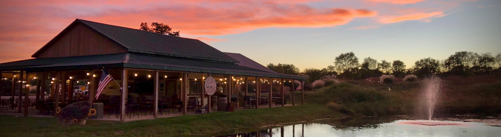 A rustic building with string lights sits beside a small pond with a fountain at sunset. The sky is vibrant with orange and pink clouds, and a U.S. flag hangs near the entrance. Trees and grass surround the peaceful scene.