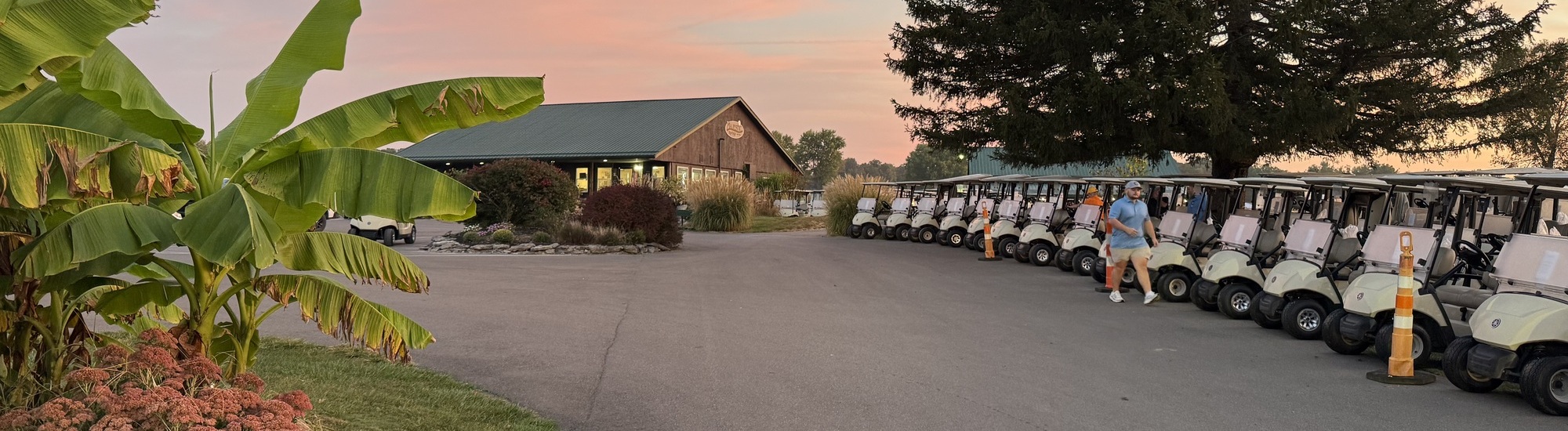 Golf carts are lined up outside a clubhouse at sunset, with a person standing near the carts. Lush plants and a large tree are visible, creating a calm and welcoming atmosphere.