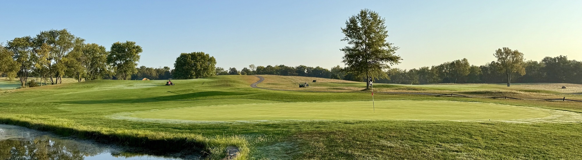 A wide view of a golf course with a manicured green, a flagstick, rolling hills, trees, and a small pond in the foreground under a clear blue sky. Golf carts and players are visible in the distance.