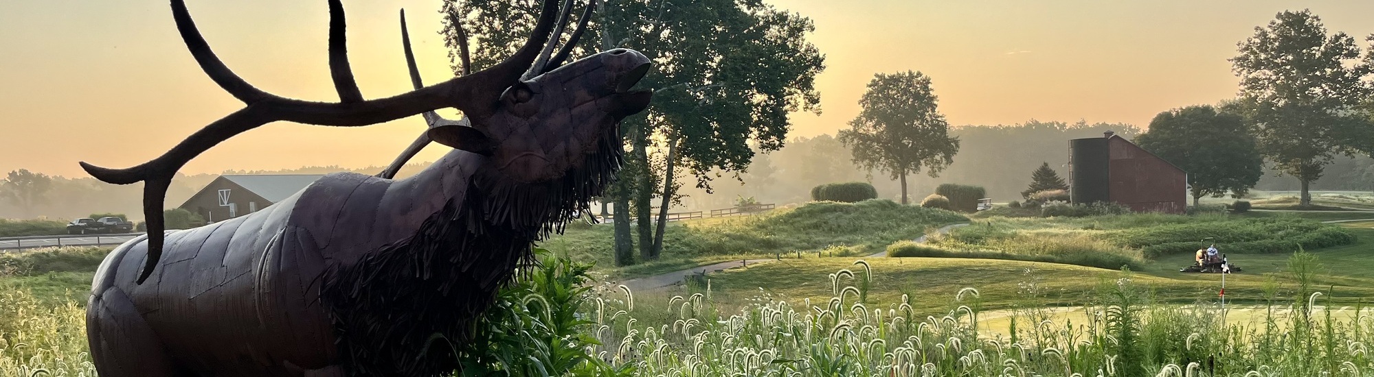 A metal elk sculpture stands in tall grass at sunrise with trees, a barn, and a person riding a lawn mower in the distance on a misty landscape.