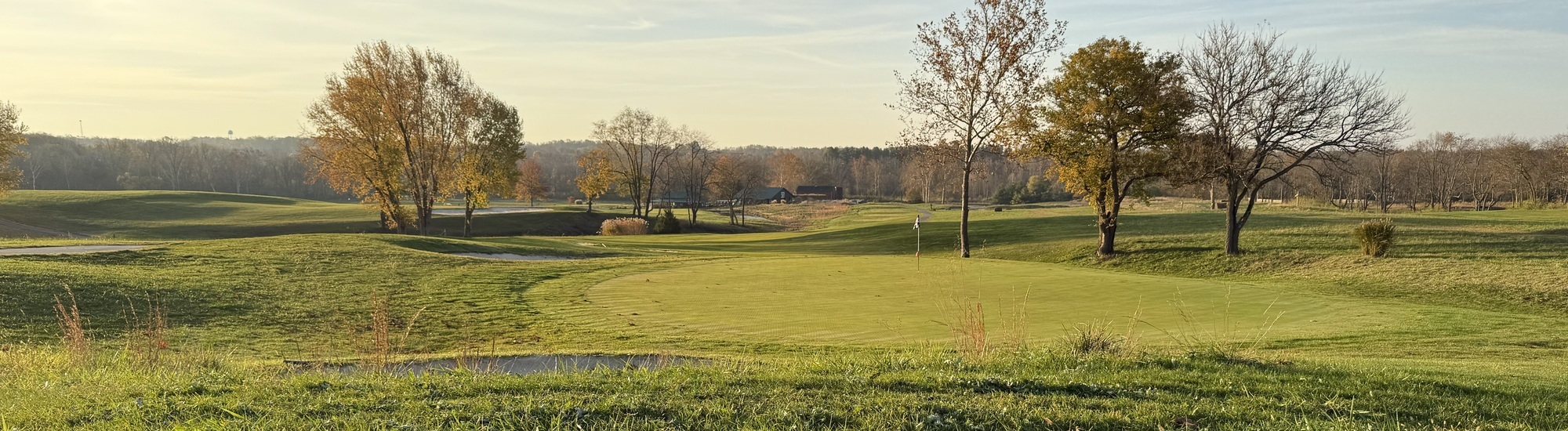 A scenic golf course with rolling green hills, sand bunkers, and scattered autumn trees under a clear sky. In the distance are more trees and a clubhouse, all bathed in soft, golden sunlight.